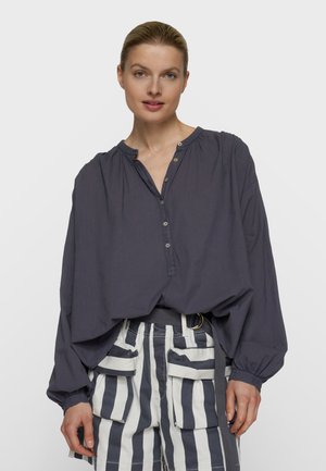 Mujer que lleva una blusa suelta de color gris oscuro con botones, mangas abullonadas y pantalones cortos de talle alto con rayas verticales en blanco y negro y grandes bolsillos.