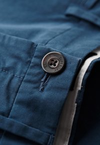 Tissu bleu avec une texture douce, présentant un bouton noir gravé "SUPERDRY". Coutures visibles et une taille soigneusement repliée.