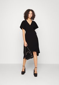 Black wrap dress with short flutter sleeves, V-neckline, high-low hem, paired with a small black bag and high-heeled platform shoes.