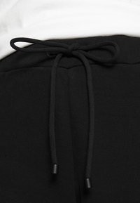 Nära bild av svarta sweatpants med en knuten dragsko i midjan, mot en vit bakgrund.