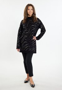 Maglione tunica nero con motivi a onde in argento, oro e bianco. Abbinato a pantaloni neri aderenti e scarpe nere lucide.