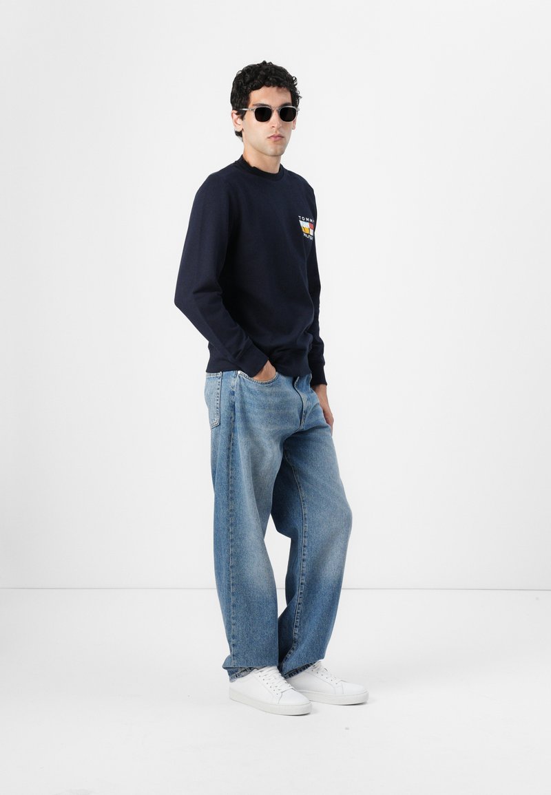 Camicia a maniche lunghe navy con logo, jeans a gamba larga blu chiaro e sneaker bianche, accompagnati da occhiali da sole rotondi neri.
