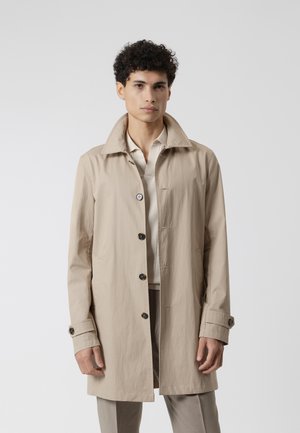 Joven con abrigo largo beige con botones, de pie frente a un fondo liso gris claro, mirando hacia adelante con expresión neutral.