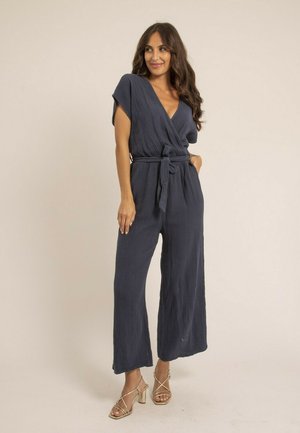 Femme debout, main dans la poche, portant une combinaison texturée bleu marine avec ceinture et des sandales à talons beiges à lanières, sur un fond beige.