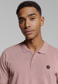 Lichtroze poloshirt van gestructureerde stof, met drie knopen en een hexagonale logo patch op de linkerkant van de borst. Korte mouwen.