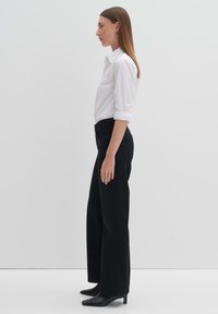 Schwarze hoch taillierte weit geschnittene Jeans mit Knopf- und Reißverschluss, kombiniert mit einem taillierten weißen Hemd mit Knöpfen. Schwarze Stiefeletten mit Absatz.