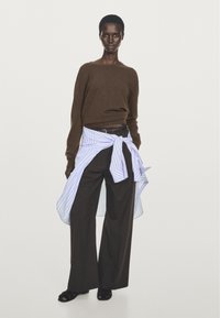 Brun stickad tröja kombinerad med vida bruna byxor, lager på lager med en blå randig skjorta knuten i midjan. Svarta sandaler med öppen tå.