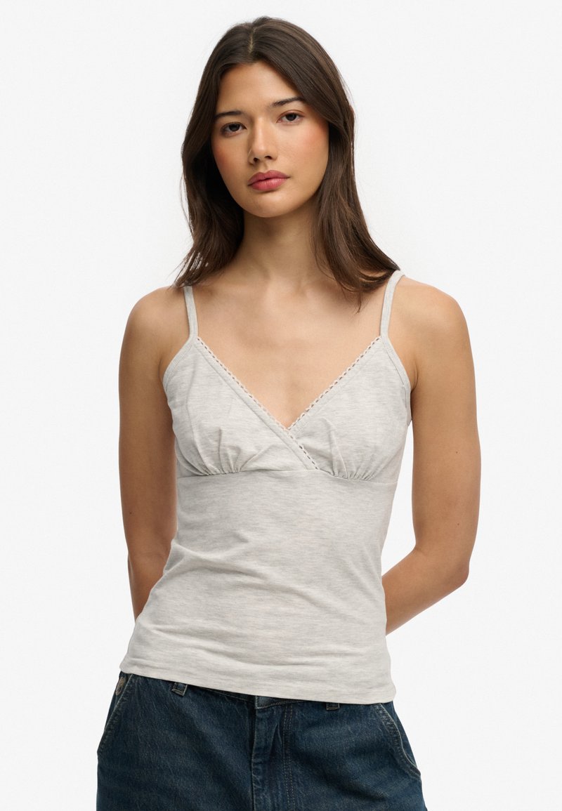 Vrouw met lang bruin haar, gekleed in een lichtgrijs mouwloos topje met V-hals en donkere spijkerbroek, tegen een effen witte achtergrond.