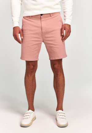 Homme portant un short rose clair, une chemise blanche à manches longues et des baskets blanches, debout sur un fond uni.