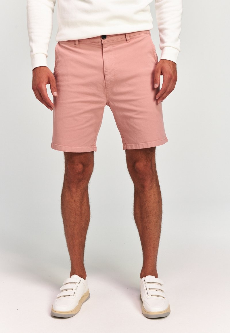 Homme portant un short rose clair, une chemise blanche à manches longues et des baskets blanches, debout sur un fond uni.