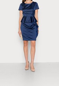 Marinblå satin peplumklänning med korta ärmar och asymmetrisk volangdetalj. Modellen håller i en liten kuvertväska och har på sig nudefärgade högklackade skor.