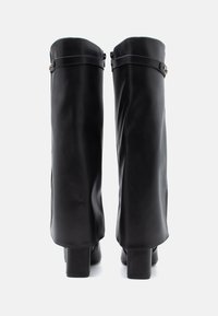 Μαύρες δερμάτινες μπότες έως το γόνατο με λεία υφή, τετράγωνα τακούνια και μία απλή λεπτομέρεια με λουρί στην κορυφή. Μινιμαλιστικός σχεδιασμός.