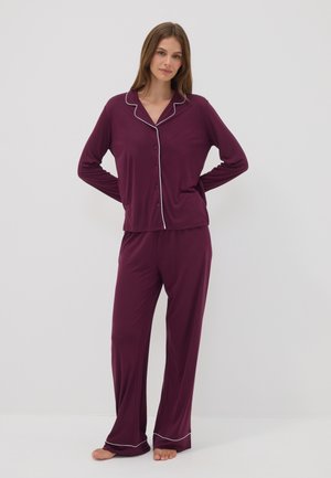 Vrouw draagt een bordeauxrode pyjama met lange mouwen, knopen en witte biezen, staat blootsvoets tegen een effen lichtgrijze achtergrond.