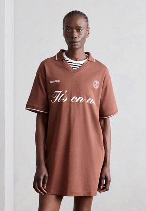 Lühikeste palmikutega naine, kes kannab pruuni üle suurusega polo-kleiti, millel on kirjas "It's on us", ja valget triibulist särki all, seisab halli seina ääres.