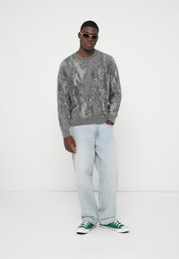 Γκρίζα υφασμάτινη ζακέτα με στρογγυλή λαιμόκοψη, ανοιχτό μπλε φαρδιά τζιν και πράσινα αθλητικά παπούτσια. Τα αξεσουάρ περιλαμβάνουν κομψά γυαλιά ηλίου.