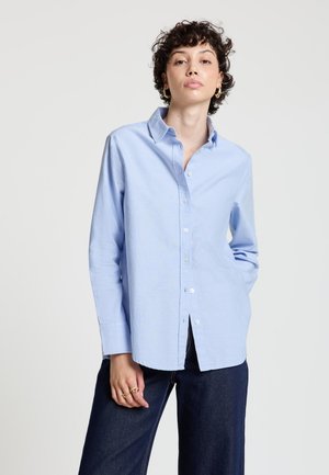 Frau mit kurzen lockigen Haaren, die ein hellblaues Hemd mit Knöpfen und dunkle Jeans trägt und vor einem schlichten Hintergrund steht.