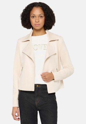 Crème-kleurig jasje van imitatiesuède met een reverskraag, ritssluitingen op de zakken en een aansluitend model, gecombineerd met een wit overhemd en donkere spijkerbroek.