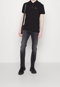 Svart zip-neck polo skjorta, slim-fit grå jeans och svarta ankelskor. Modellen bär en svart axelremsväska. Enkel design, avslappnad stil.