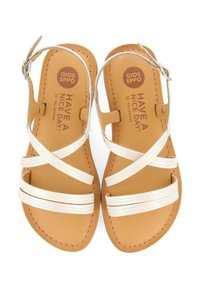 Sandalias de cuero blancas con tiras entrelazadas sobre una plantilla de cuero marrón. Forma redondeada, suela plana y detalle de correa ajustable en el tobillo.