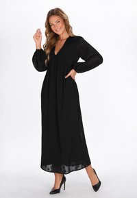 Rochie maxi neagră cu decolteu adânc în V, mâneci lungi și bufante, dintr-un material texturat. Asortată cu pantofi cu toc înalt negri.