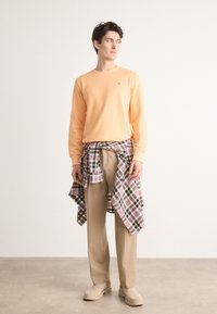Orange crewneck sweatshirt, beige pants, and plaid shirt tied at the waist. Smooth fabric, relaxed fit, and neutral footwear.
