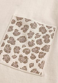 Beżowa tkanina z kwadratową łatą o wzorze w panterkę, wykonaną z brązowych i różowych cyrkonii, tworzących teksturowany akcent.