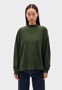 Sweatshirt vert oversize avec un col montant, des manches lâches, une texture douce et des coutures minimales, associé à un jean bleu foncé.