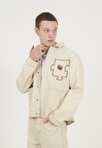 Beige farmerdzseki gombos elülső résszel, mellzseb�ével, amely egy puzzle darab mintáját viseli, és kontrasztos varrással. A modell az állára támasztott kézzel pózol.
