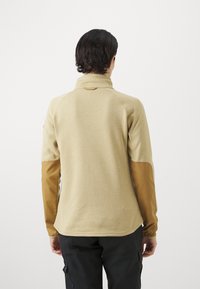 Pullover in felpa beige con maniche in pelle marrone, caratterizzato da un colletto alto, una texture morbida e un orlo posteriore più lungo.