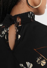 Gros plan d'une personne portant un haut noir à motifs floraux avec un col montant froncé fermé par des boutons et une grande boucle d'oreille en or.