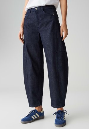 Person trägt lockere dunkelblaue Jeanshosen und blaue Turnschuhe mit weißen Streifen, steht auf grauem Hintergrund.