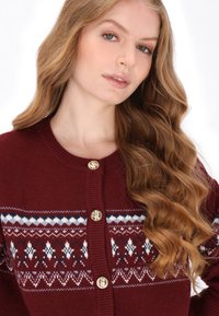 Bordowy dzianinowy kardigan z okrągłym dekoltem, ozdobiony geometrycznym wzorem w białe i niebieskie kolory oraz złotymi guzikami z przodu.