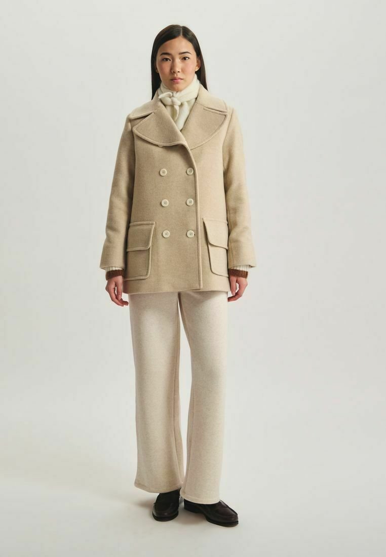 Manteau en laine beige avec un design croisé, des revers larges et deux poches avant. Associé à un pantalon large crème et des chaussures noires.