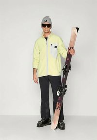 Veste en polaire jaune avec des accents gris, pantalon de ski noir, bonnet gris et lunettes de soleil, tenant un ski avec fixations et marque visible.