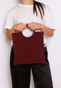 Borsa a spalla bordeaux con manico circolare, texture morbida e forma rettangolare minimalista. Indossata da una persona con una maglietta bianca e pantaloni neri.