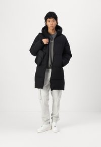Abrigo de plumas negro con capucha, con zipper frontal, dos bolsillos laterales y un diseño de longitud media, usado sobre una camiseta gris y pantalones claros.
