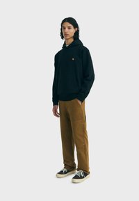 Felpa nera con cappuccio e tasca frontale, abbinata a pantaloni cargo beige con cerniere laterali e bottoni metallici. Sneakers nere con lacci bianchi.