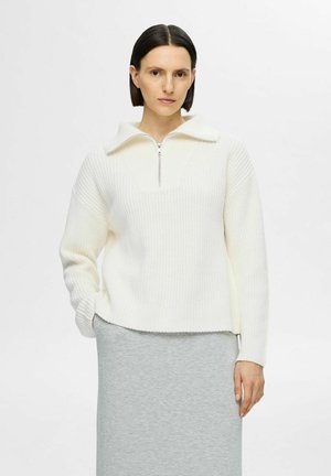 Pullover bianco in maglia con colletto a zip quarti, motivo testurizzato e design con spalla cadente. Indossato con una gonna grigia.