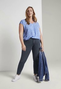 Hellblaue Kurzarmbluse mit weißem Blumenmuster, kombiniert mit dunklen Skinny-Jeans und weißen Sneakern; eine Jeansjacke wird in der rechten Hand gehalten.