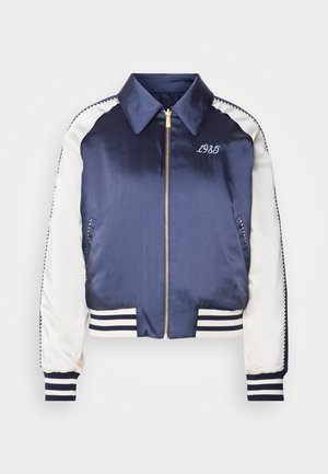 Casaco bomber de satino azul marinho com mangas brancas, punhos e bainha canelados, fecho dourado e bordado "1985" no peito.