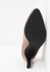Chaussure à talon haut en daim marron à bout pointu reposant sur le côté, montrant la semelle noire texturée et le talon fin sur fond blanc.