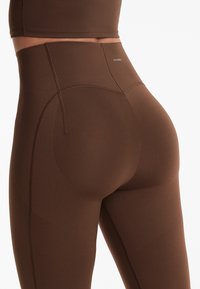 Braune hoch taillierte Leggings mit glatter Textur, die konturierte Nähte und dezentes Branding am Bund aufweisen.