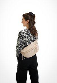 Beige Umhängetasche mit zwei Reißverschlussfächern und Lederquasten, getragen über einem schwarz-weißen gemusterten Oberteil und schwarzen Hosen.