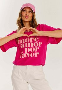 Svetle růžové oversized tričko s výrazným béžovým nápisem "more amor por favor." V kombinaci s bílými kalhotami a světle růžovou čepicí.
