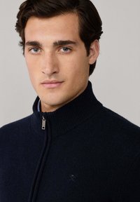 Marineblauer Reißverschluss-Pullover mit hohem Kragen, aus weichem Material gefertigt, mit einem dezenten Logo auf der Brust und einer strukturierten Oberfläche.
