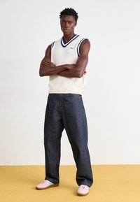 Pull sans manches en maille crème avec des accents bleu marine, associé à un jean bleu foncé à coupe large et des chaussures roses avec des accents vert menthe.