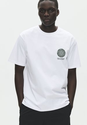 Osoba nosiaca jednoduché biele tričko s malým kruhovým logom a textom na ľavej strane hrudníka, stojaca s rukami v vreckách.