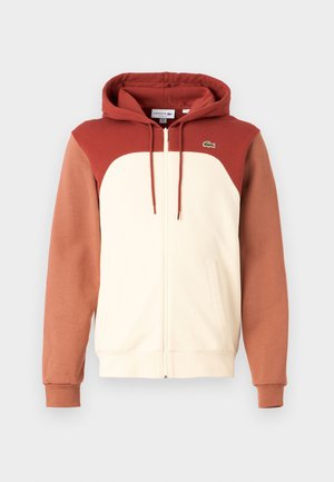 Sudadera con capucha y cremallera que presenta un frente crema, hombros y mangas en color óxido, con un pequeño logotipo verde en el pecho. Textura de tela suave.