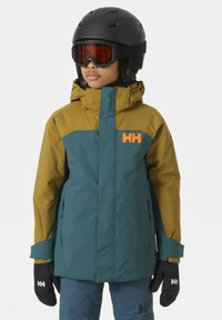 Skijacke in Grün und Senfgelb, mit hohem Kragen, Logo auf der Brust und Reißverschlusstaschen. Mattes Finish, kombiniert mit schwarzen Handschuhen.