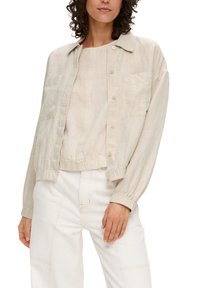 Light beige linen jacket with a collared neck, button front, two chest pockets, and elastic cuffs, paired with a similar-tone top and white pants.